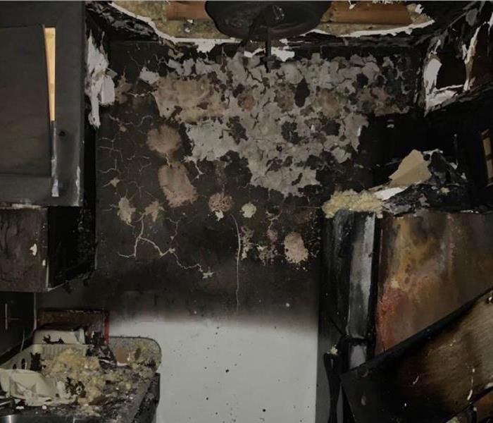 Photo of a wall with burnt markings and fire damage. Next to the wall you see a burnt fridge as well as burnt cabinets.